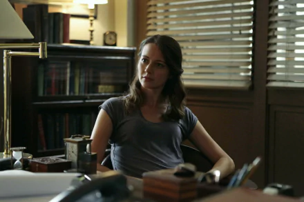 A close-up of Amy Acker from the TV show *Person of Interest*, seated at a desk in a warmly lit room, exuding a contemplative expression amidst books and office items.