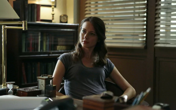 A close-up of Amy Acker from the TV show *Person of Interest*, seated at a desk in a warmly lit room, exuding a contemplative expression amidst books and office items.