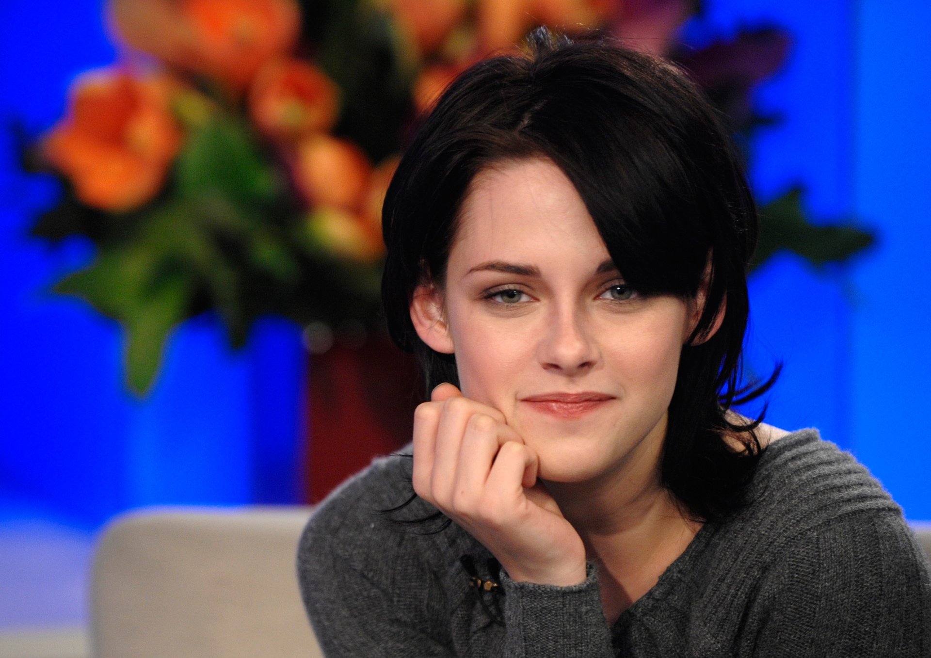 A close-up of Kristen Stewart, smiling thoughtfully with her chin resting on her hand, set against a blurred background of colorful flowers. An elegant HD desktop wallpaper.