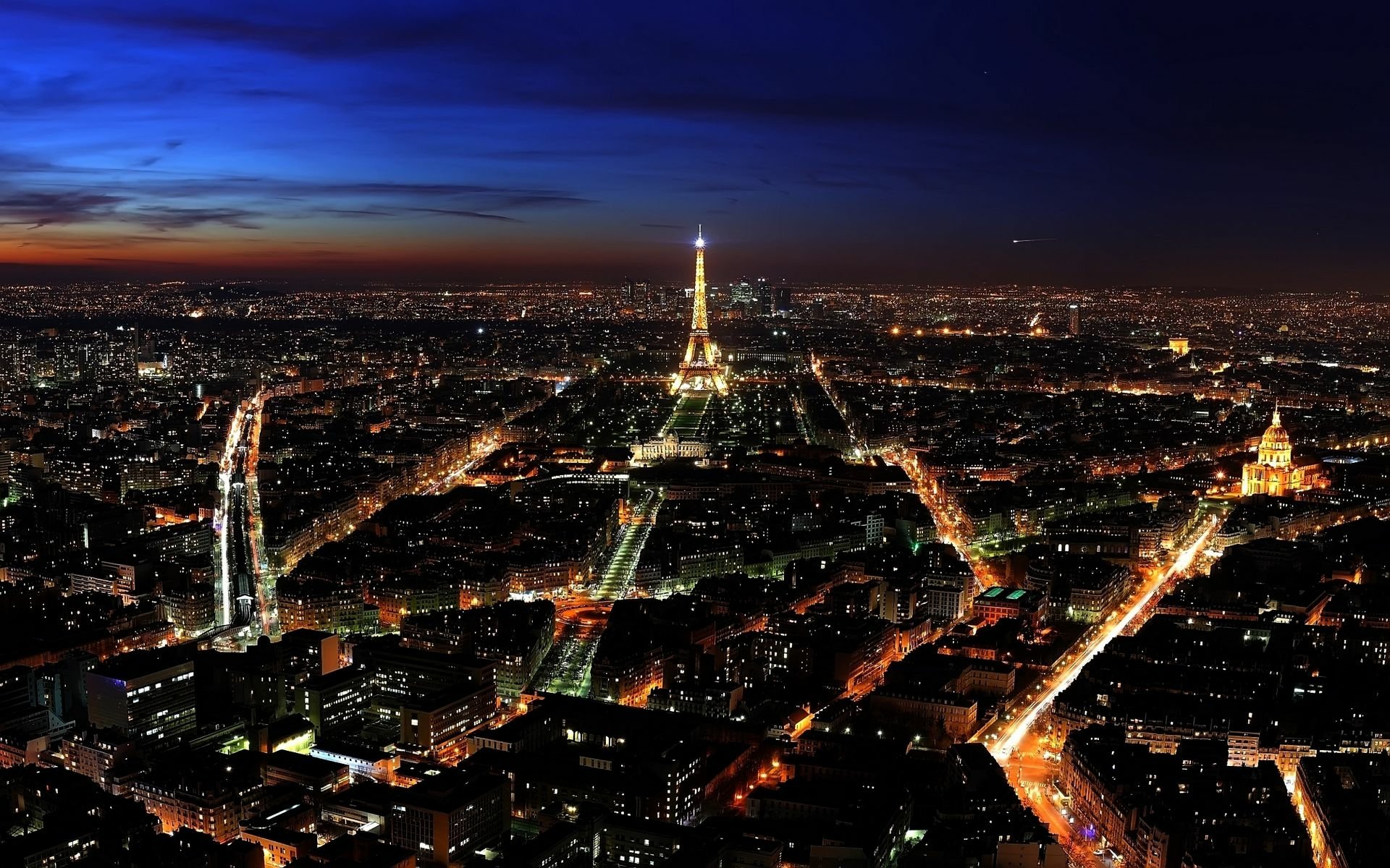 HD desktop wallpaper showcasing a stunning nighttime aerial view of Paris with the illuminated Eiffel Tower at the center against a deep blue sky.