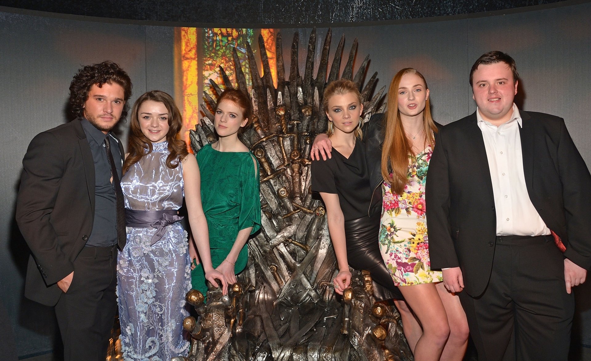 A group of popular actors, including Maisie Williams, Kit Harington, and Sophie Turner, poses together near the Iron Throne, showcasing their connection to the iconic series.