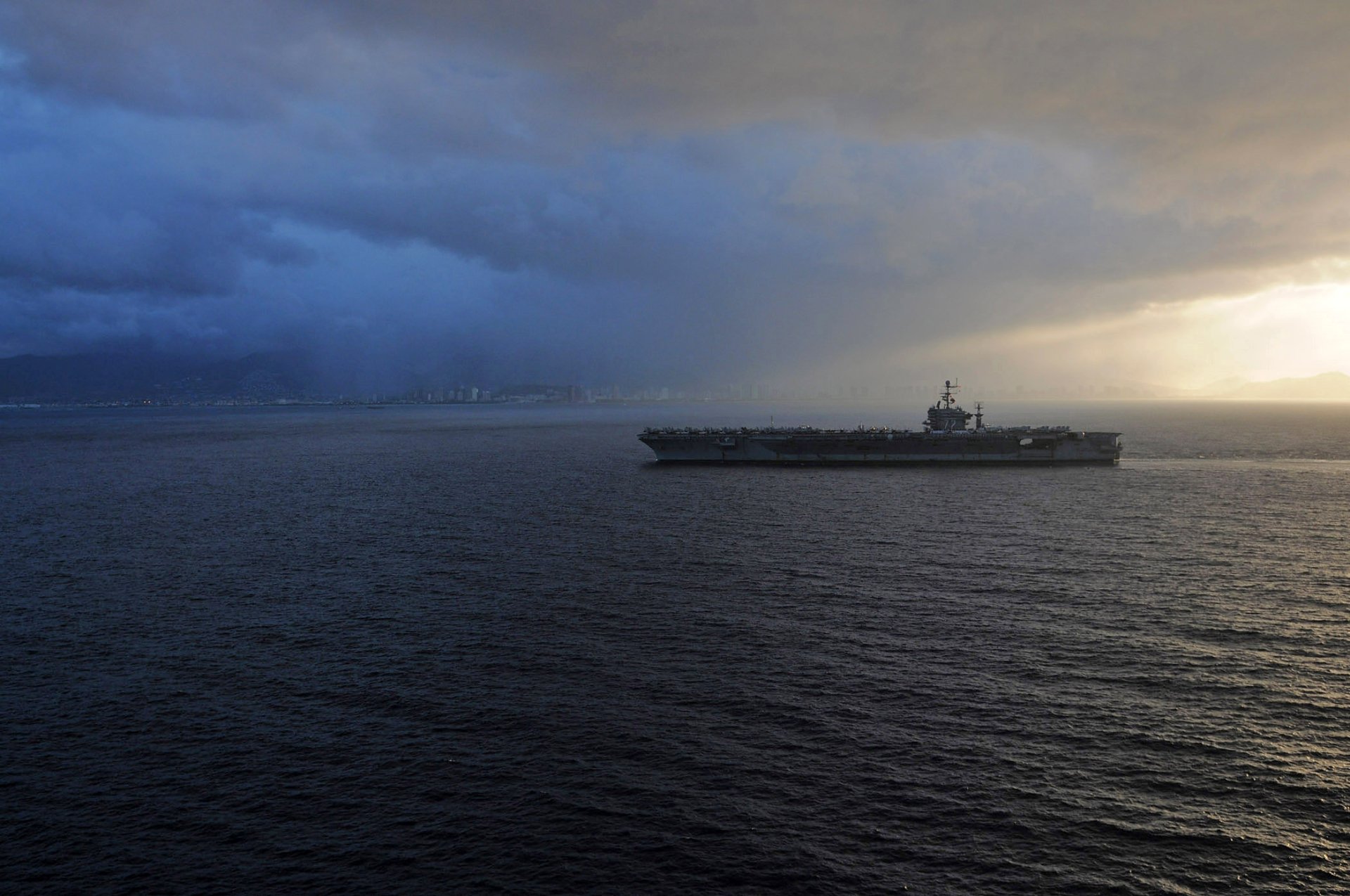 HD desktop wallpaper featuring the USS Abraham Lincoln (CVN-72) aircraft carrier sailing through calm waters under a dramatic cloudy sky.