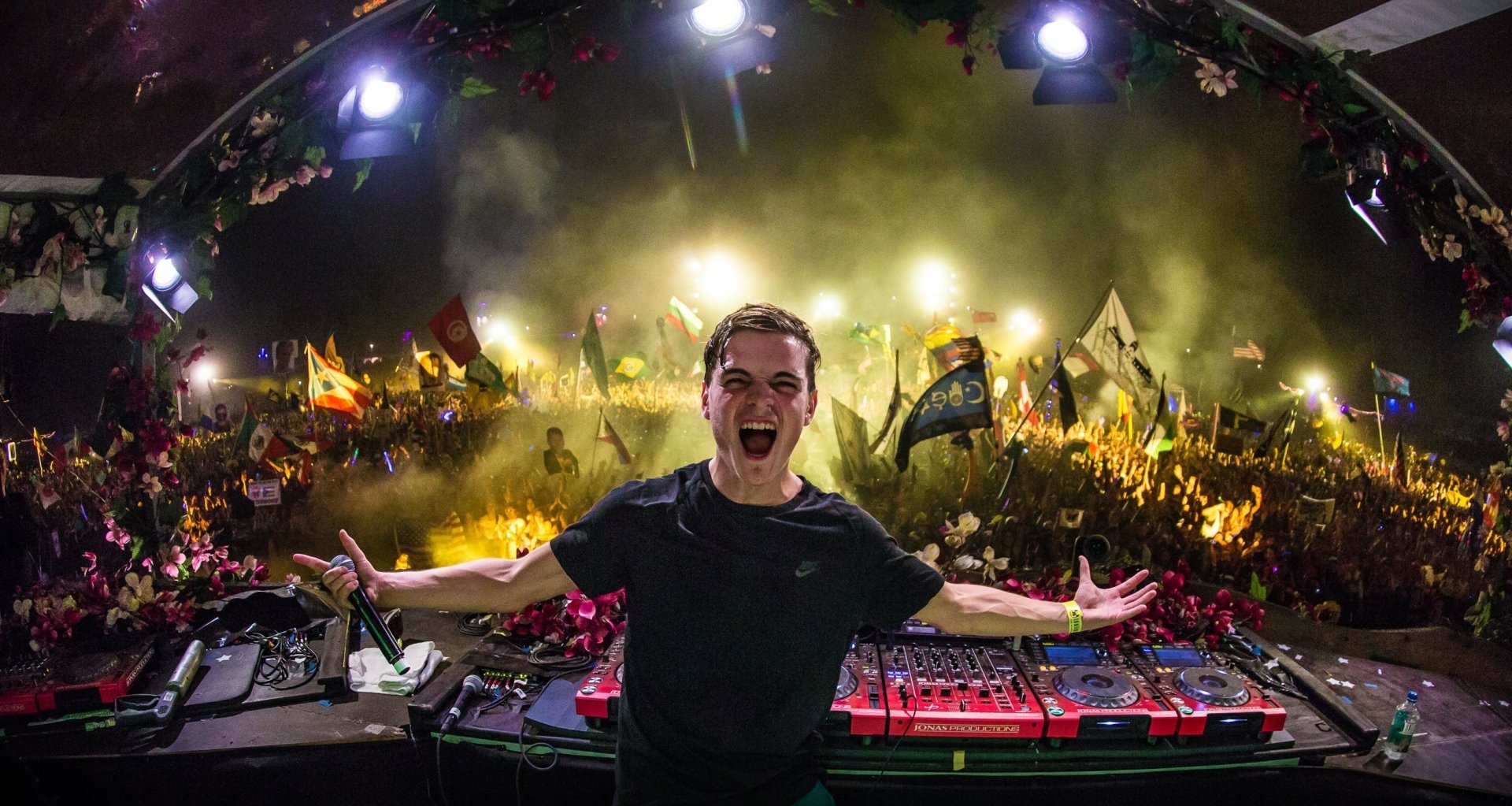 HD wallpaper featuring Martin Garrix enthusiastically performing on stage in front of a large, energetic crowd with bright lights and vibrant flags.