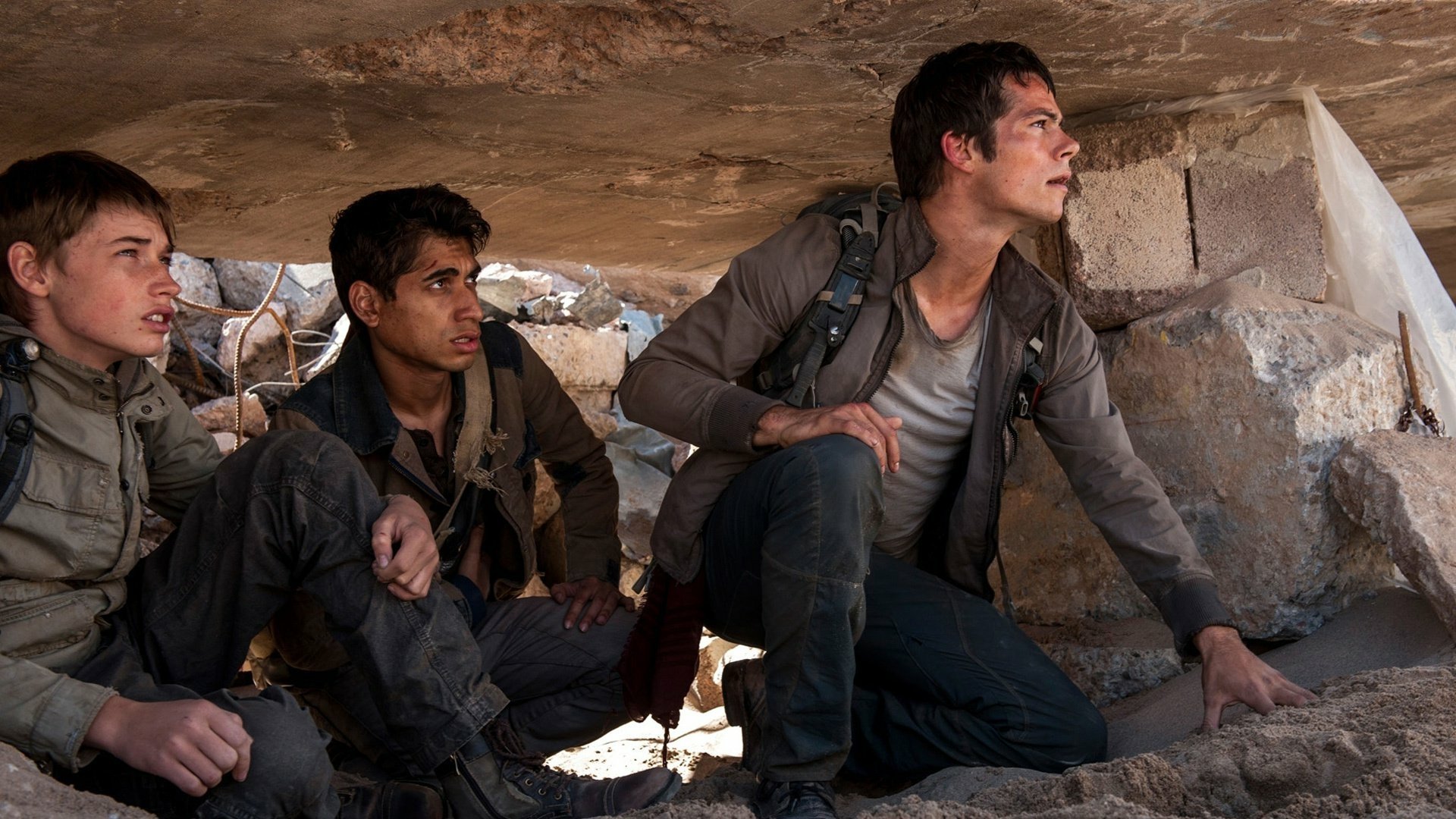 HD PC desktop wallpaper: scene from Maze Runner: The Scorch Trials — three young men crouch beneath rubble in a sunlit desert ruin.