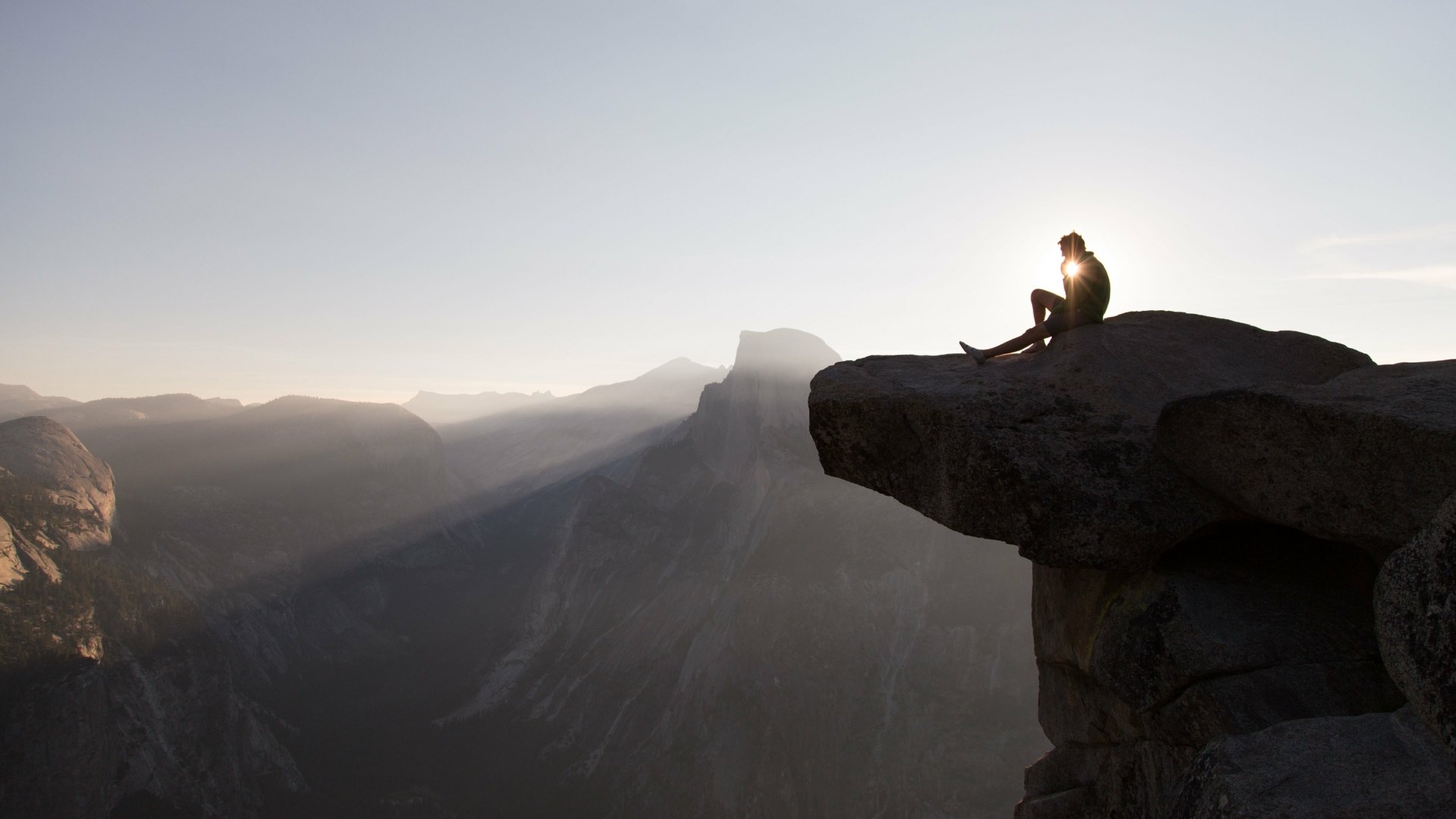 A person sits on a mountain edge bathed in a sunbeam, overlooking a vast nature landscape captured in 4K Ultra HD.