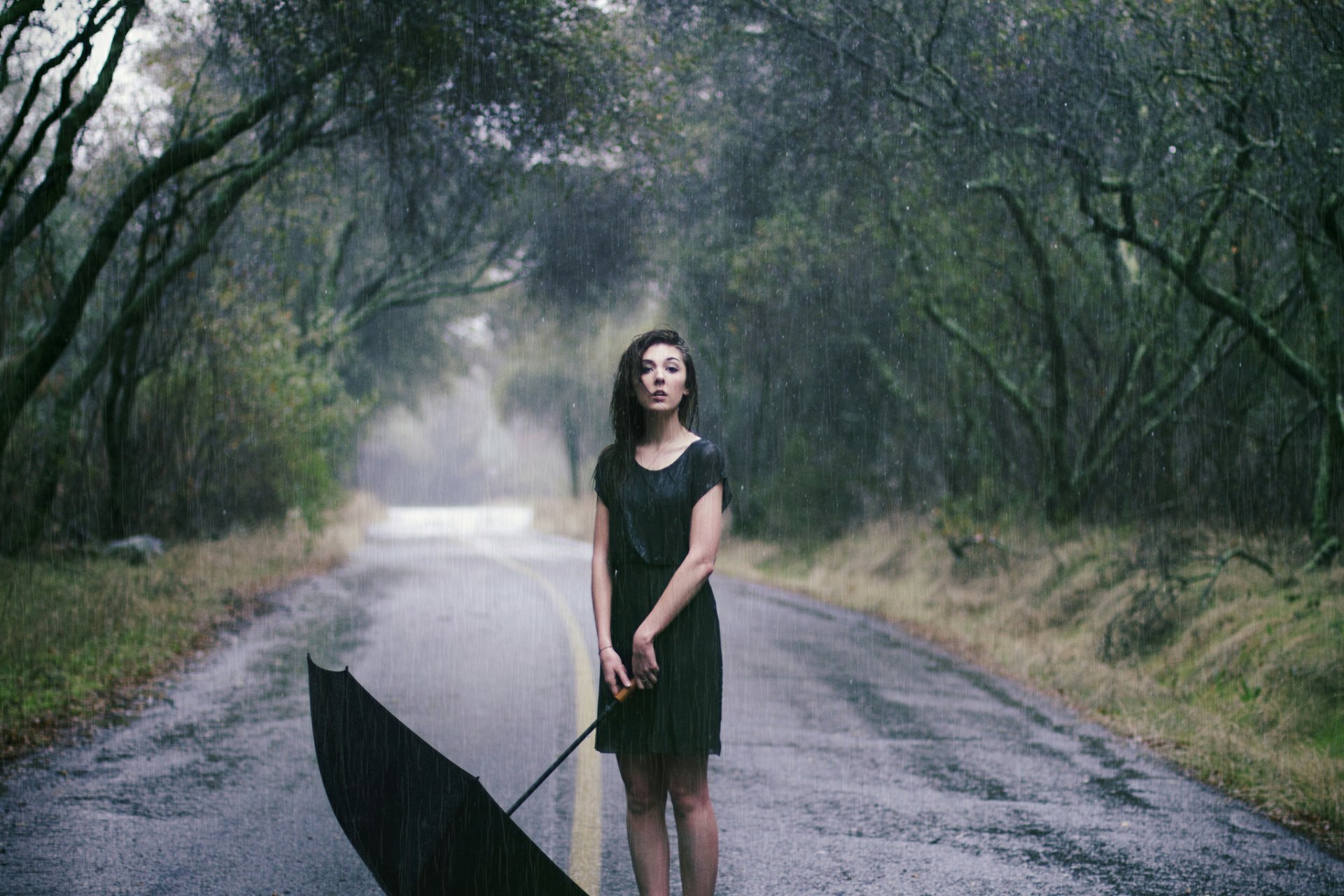 A brunette woman in a black dress stands on a rain-soaked road holding a closed umbrella, surrounded by misty, tree-lined scenery, evoking a moody atmosphere.