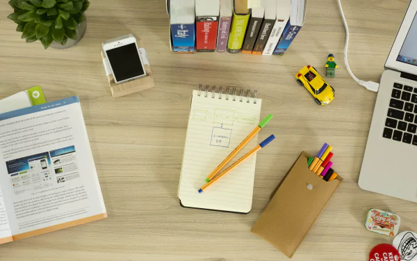 A 4K Ultra HD desktop wallpaper featuring a still life view of a desk with books, a notebook with drawings, pencils, a smartphone, and a laptop.