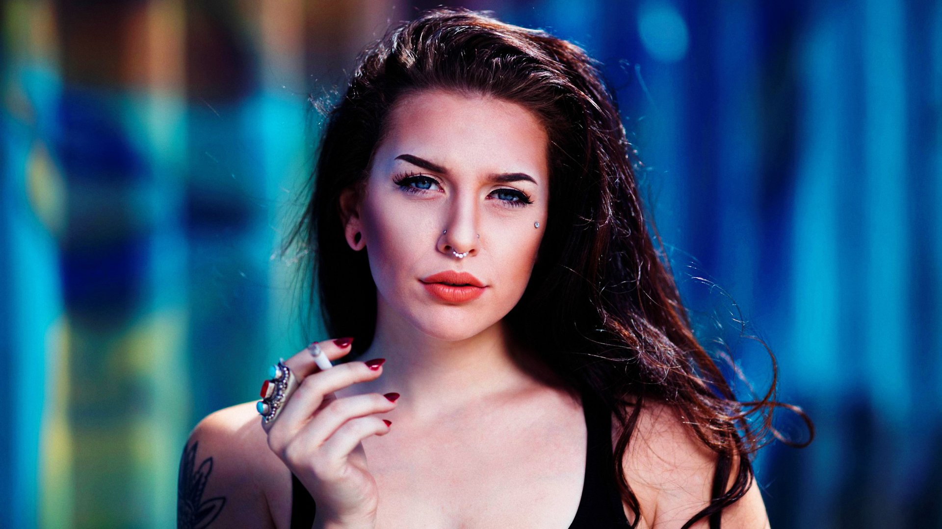 A brunette woman with blue eyes and tattoos holds a cigarette, exuding confidence against a colorful, blurred background in this striking HD desktop wallpaper.