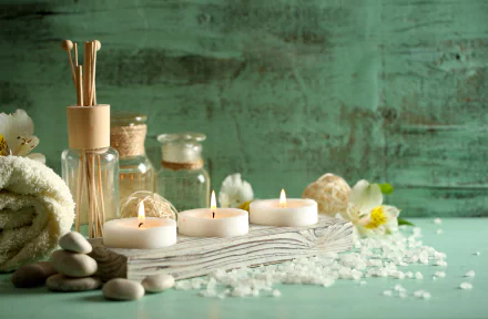 A serene spa setting features candles, stones, salt, and essential oils on a textured surface, complemented by a soft towel and natural elements, creating a relaxing atmosphere.