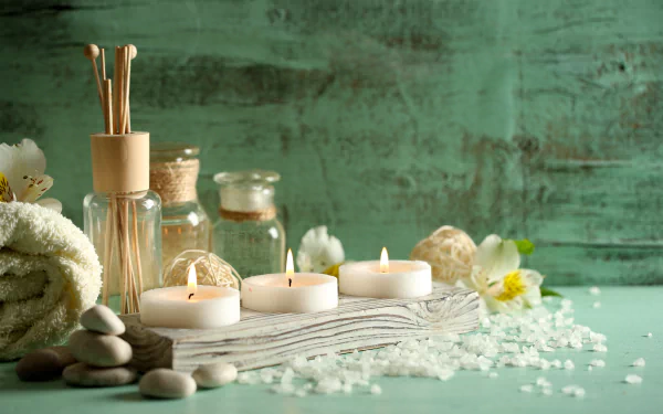A serene spa setting features candles, stones, salt, and essential oils on a textured surface, complemented by a soft towel and natural elements, creating a relaxing atmosphere.
