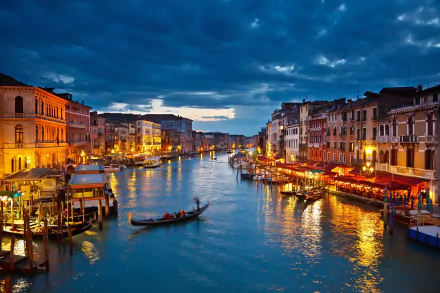 A scenic view of a Venice canal at night, illuminated by city lights, with a gondola gliding through the water. The image is a high-definition desktop wallpaper.
