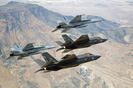 A formation of U.S. Navy jet fighters, featuring a Lockheed Martin F-35 Lightning II and Boeing F/A-18E/F Super Hornets, flying over mountainous terrain.