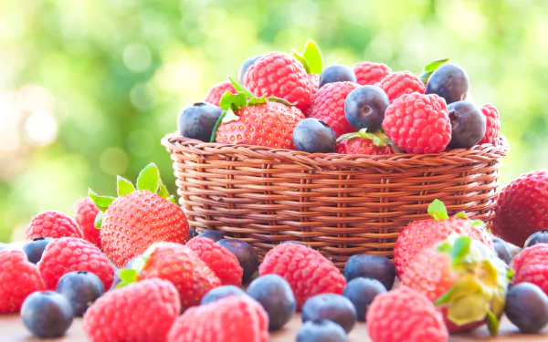 A wicker basket filled with fresh strawberries, blueberries, and raspberries sits surrounded by more berries, set against a softly blurred green background. 4K Ultra HD wallpaper.