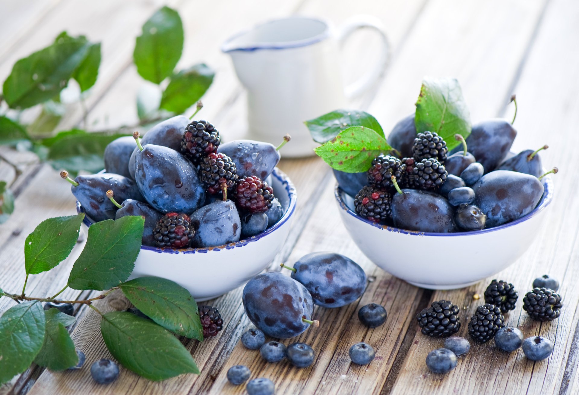 HD desktop wallpaper featuring fresh blackberries, blueberries, and plums arranged in white bowls on a wooden surface with green leaves and a blurred background.