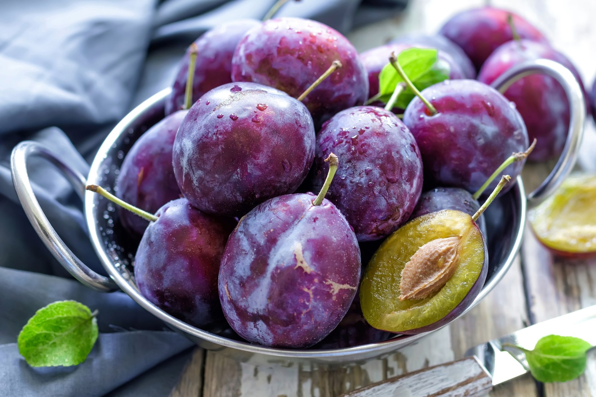 A bowl of fresh, ripe plums with a rich purple hue, highlighted by a halved plum revealing its pit. This vibrant image serves as a stunning 4K Ultra HD desktop wallpaper.