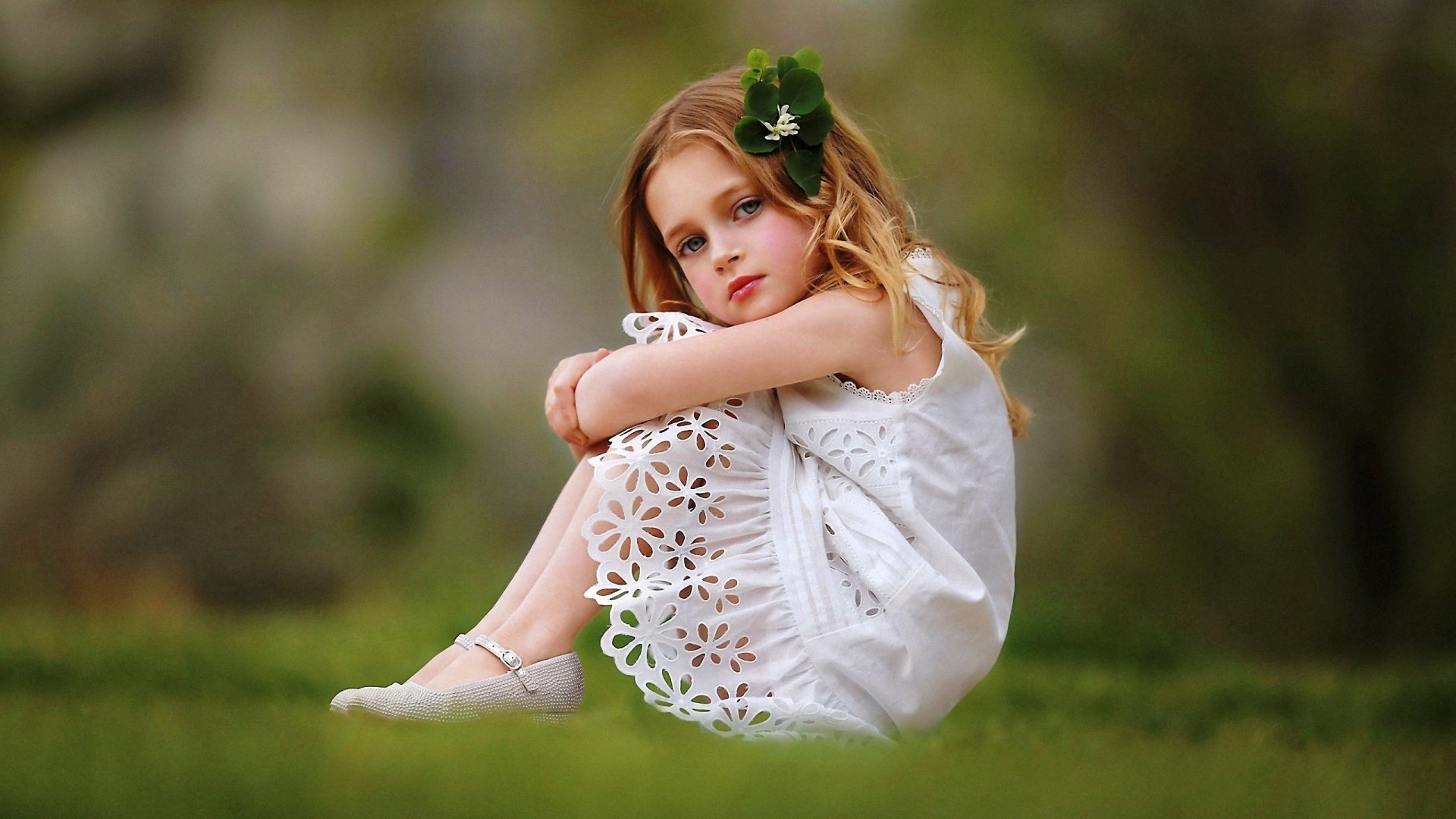 HD photography wallpaper of a young girl in a white dress sitting on grass, with a green flower in her hair and a blurred natural background.