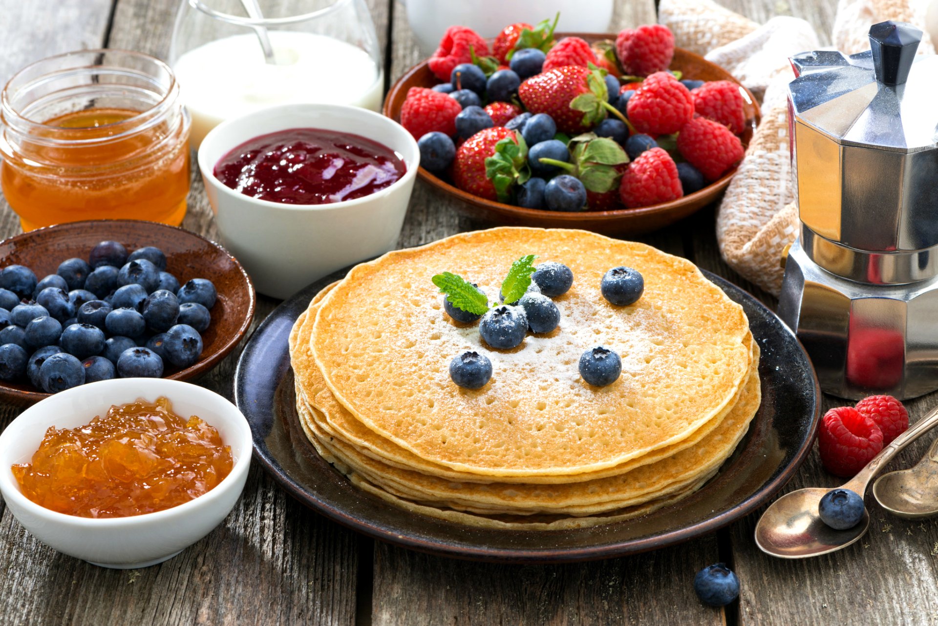 A delicious assortment of crêpes topped with blueberries, surrounded by fresh strawberries, raspberries, jars of jam, honey, and a coffee pot, creating a vibrant breakfast scene.