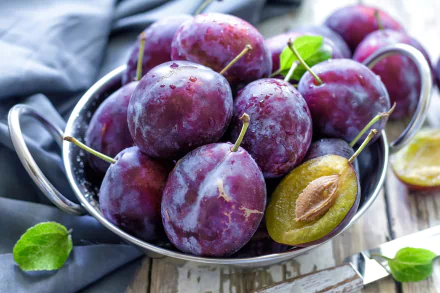 A bowl of fresh, ripe plums with a rich purple hue, highlighted by a halved plum revealing its pit. This vibrant image serves as a stunning 4K Ultra HD desktop wallpaper.
