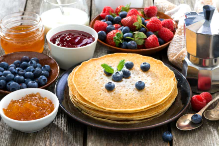 A delicious assortment of crêpes topped with blueberries, surrounded by fresh strawberries, raspberries, jars of jam, honey, and a coffee pot, creating a vibrant breakfast scene.