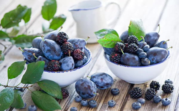 HD desktop wallpaper featuring fresh blackberries, blueberries, and plums arranged in white bowls on a wooden surface with green leaves and a blurred background.