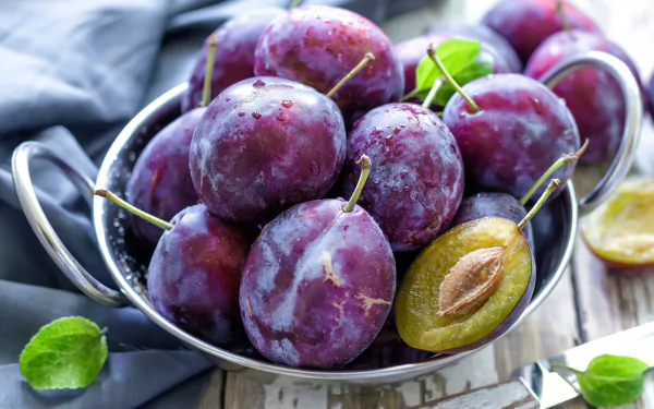 A bowl of fresh, ripe plums with a rich purple hue, highlighted by a halved plum revealing its pit. This vibrant image serves as a stunning 4K Ultra HD desktop wallpaper.