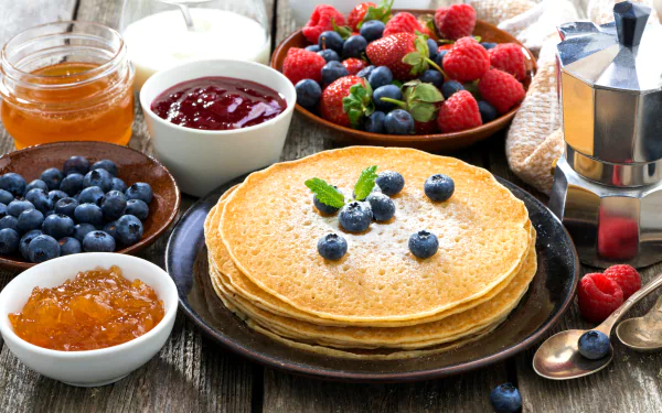 A delicious assortment of crêpes topped with blueberries, surrounded by fresh strawberries, raspberries, jars of jam, honey, and a coffee pot, creating a vibrant breakfast scene.