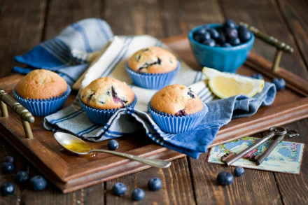 A rustic wooden tray holds freshly baked blueberry muffins in blue liners, accompanied by a bowl of blueberries and lemon slices, creating a warm, inviting food-themed wallpaper.