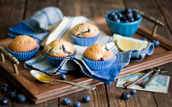 A rustic wooden tray holds freshly baked blueberry muffins in blue liners, accompanied by a bowl of blueberries and lemon slices, creating a warm, inviting food-themed wallpaper.