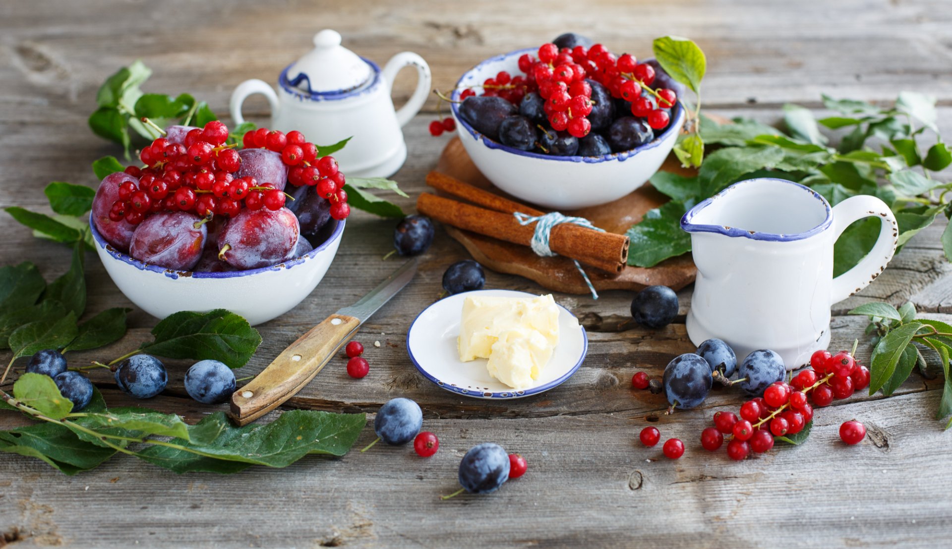 A vibrant HD desktop wallpaper featuring bowls of fresh plums, currants, and blueberries arranged on a rustic wooden table with cream, butter, and greenery.