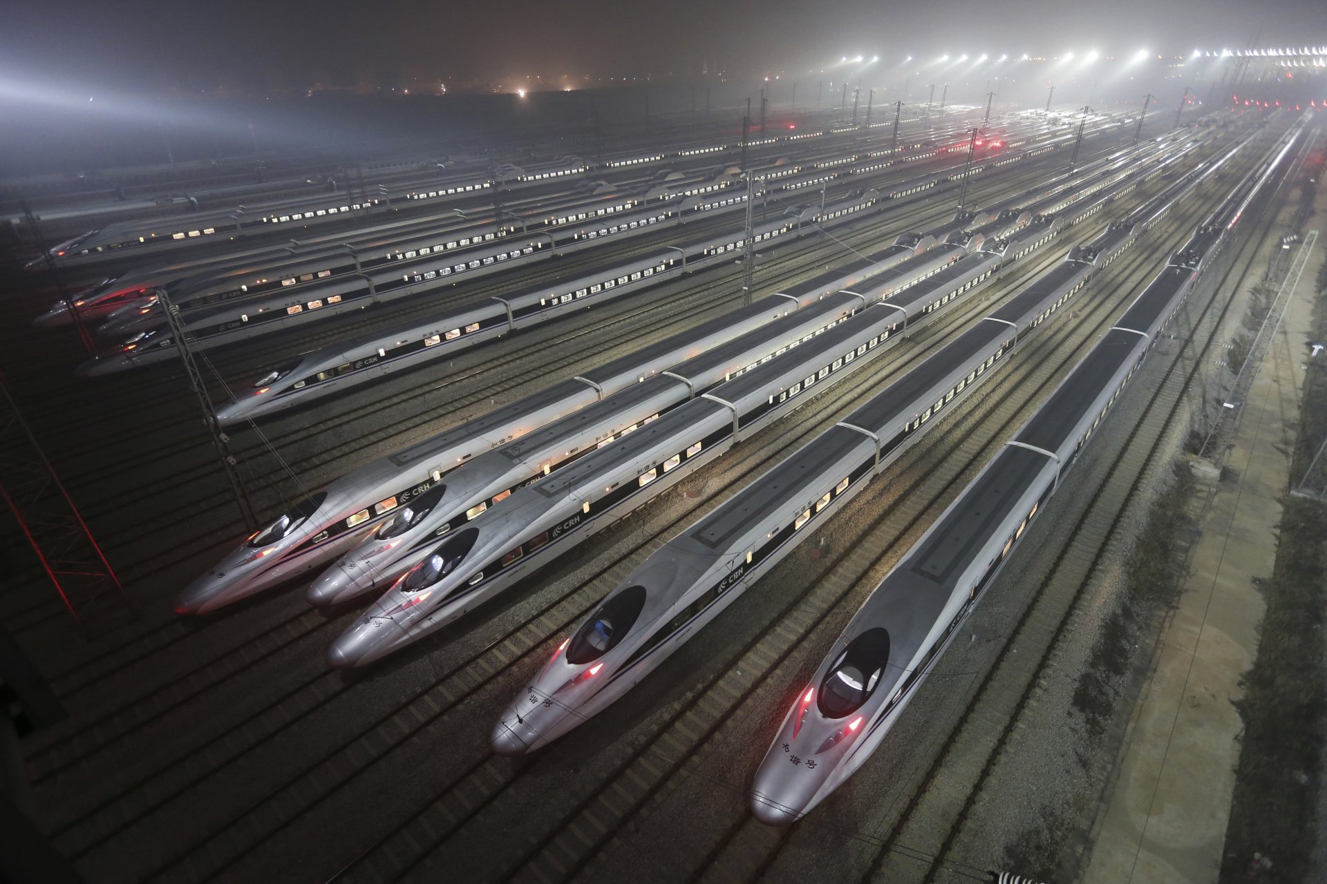 HD desktop wallpaper showing multiple sleek high-speed trains parked on parallel tracks under bright lights at night in a large rail yard.