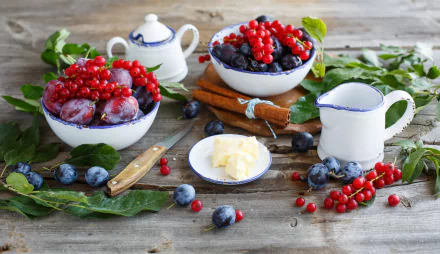 A vibrant HD desktop wallpaper featuring bowls of fresh plums, currants, and blueberries arranged on a rustic wooden table with cream, butter, and greenery.