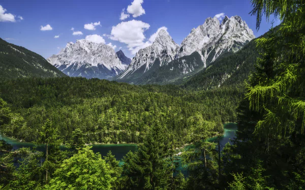 Panoramic HD desktop wallpaper of a river winding through dense forest with majestic Bavarian Alps mountains under a bright blue sky in Germany's natural landscape.