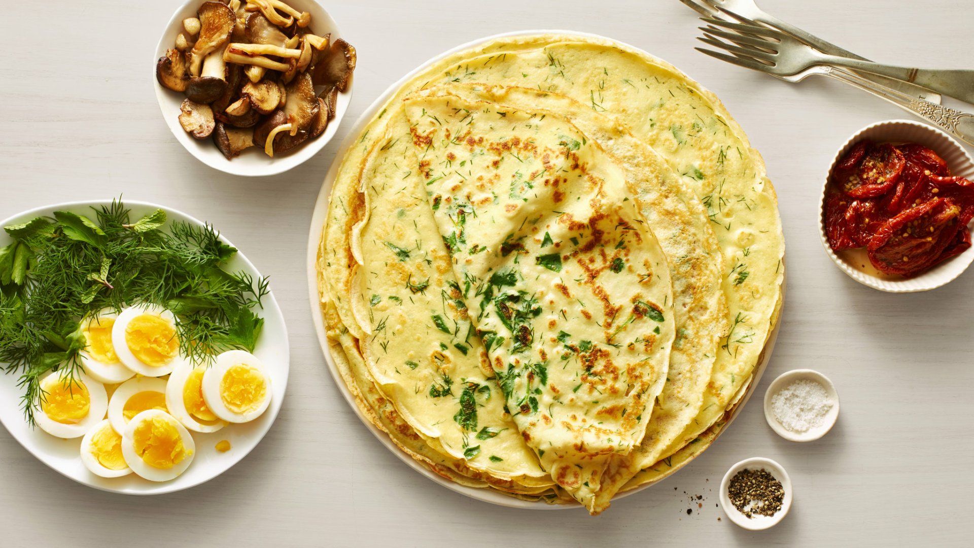 HD PC desktop wallpaper of food: savory crêpes on a plate garnished with herbs, surrounded by bowls of boiled eggs, mushrooms, sun-dried tomatoes and seasonings.