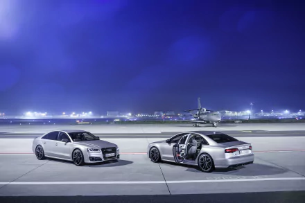 Two silver Audi S8 cars parked on an airport runway at night with an airplane and terminal lights in the background, captured in 4K Ultra HD.
