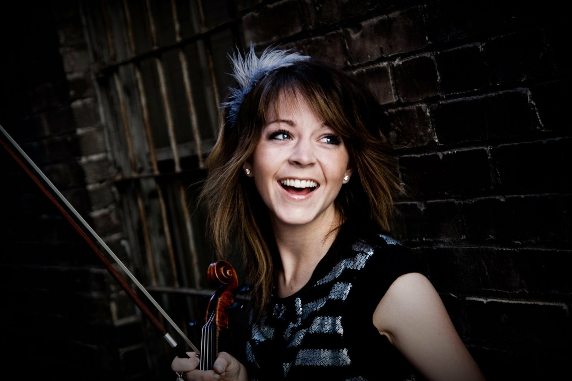 HD desktop wallpaper of musician Lindsey Stirling smiling while holding her violin against a dark brick wall background.