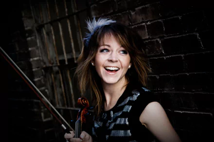HD desktop wallpaper of musician Lindsey Stirling smiling while holding her violin against a dark brick wall background.