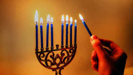 A hand lighting the shamash candle on a menorah with eight blue candles lit, captured in 4K Ultra HD for a vivid Hanukkah holiday desktop wallpaper.