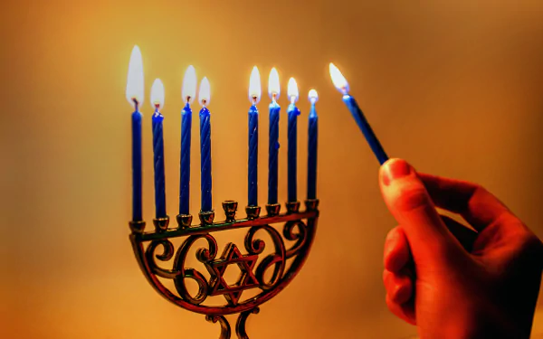 A hand lighting the shamash candle on a menorah with eight blue candles lit, captured in 4K Ultra HD for a vivid Hanukkah holiday desktop wallpaper.