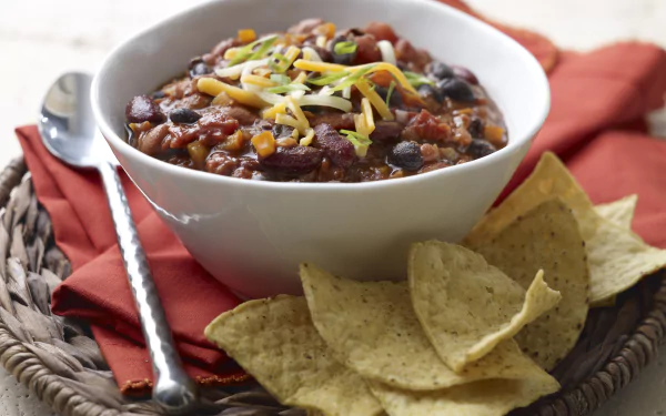 5K Ultra HD PC desktop wallpaper/background: hearty bowl of chili with beans, corn and shredded cheese, served with tortilla chips on a woven placemat and red napkin.