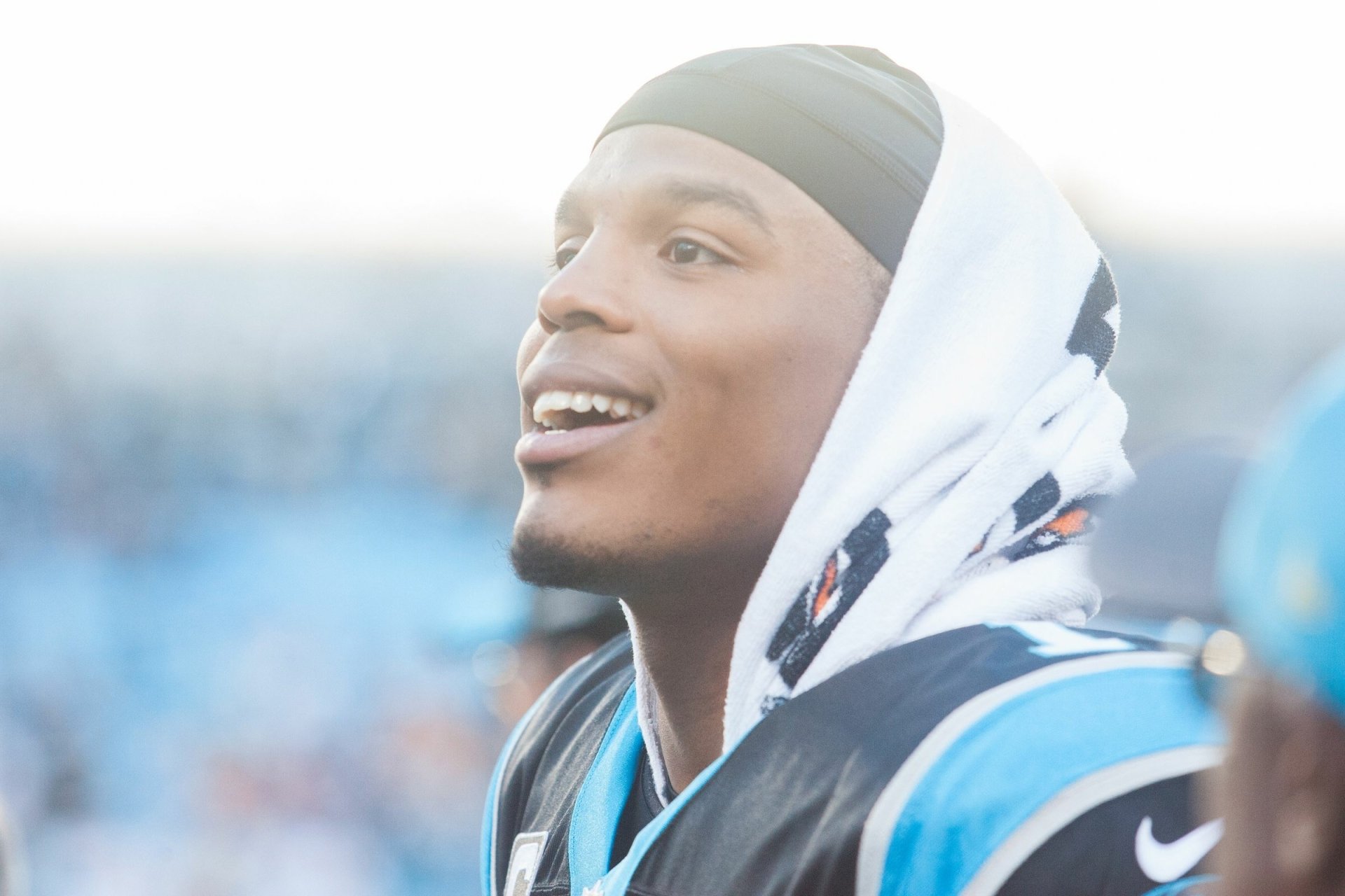 HD PC desktop wallpaper: close-up of a smiling Carolina Panthers football player in uniform with a hooded towel, bright stadium lights and blurred crowd behind him.