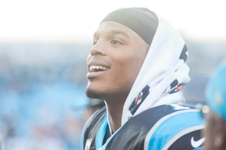 HD PC desktop wallpaper: close-up of a smiling Carolina Panthers football player in uniform with a hooded towel, bright stadium lights and blurred crowd behind him.