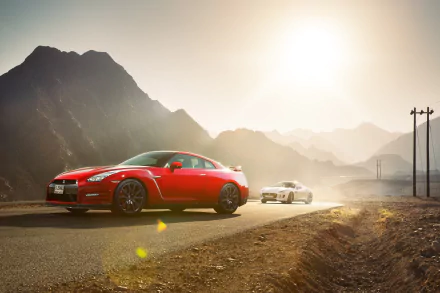A striking HD desktop wallpaper featuring a Nissan GT-R and a Jaguar F-Type driving along a mountain road, illuminated by the warm glow of the sun.