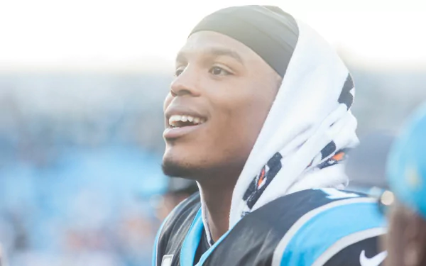 HD PC desktop wallpaper: close-up of a smiling Carolina Panthers football player in uniform with a hooded towel, bright stadium lights and blurred crowd behind him.