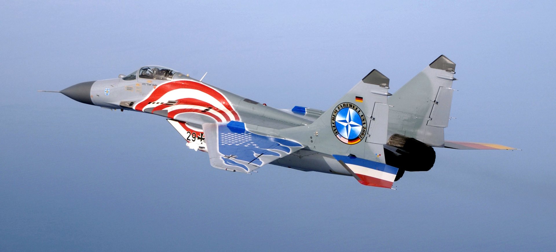 HD desktop wallpaper featuring a military Mikoyan MiG-29 fighter jet in flight with patriotic red, white, and blue markings against a clear blue sky background.