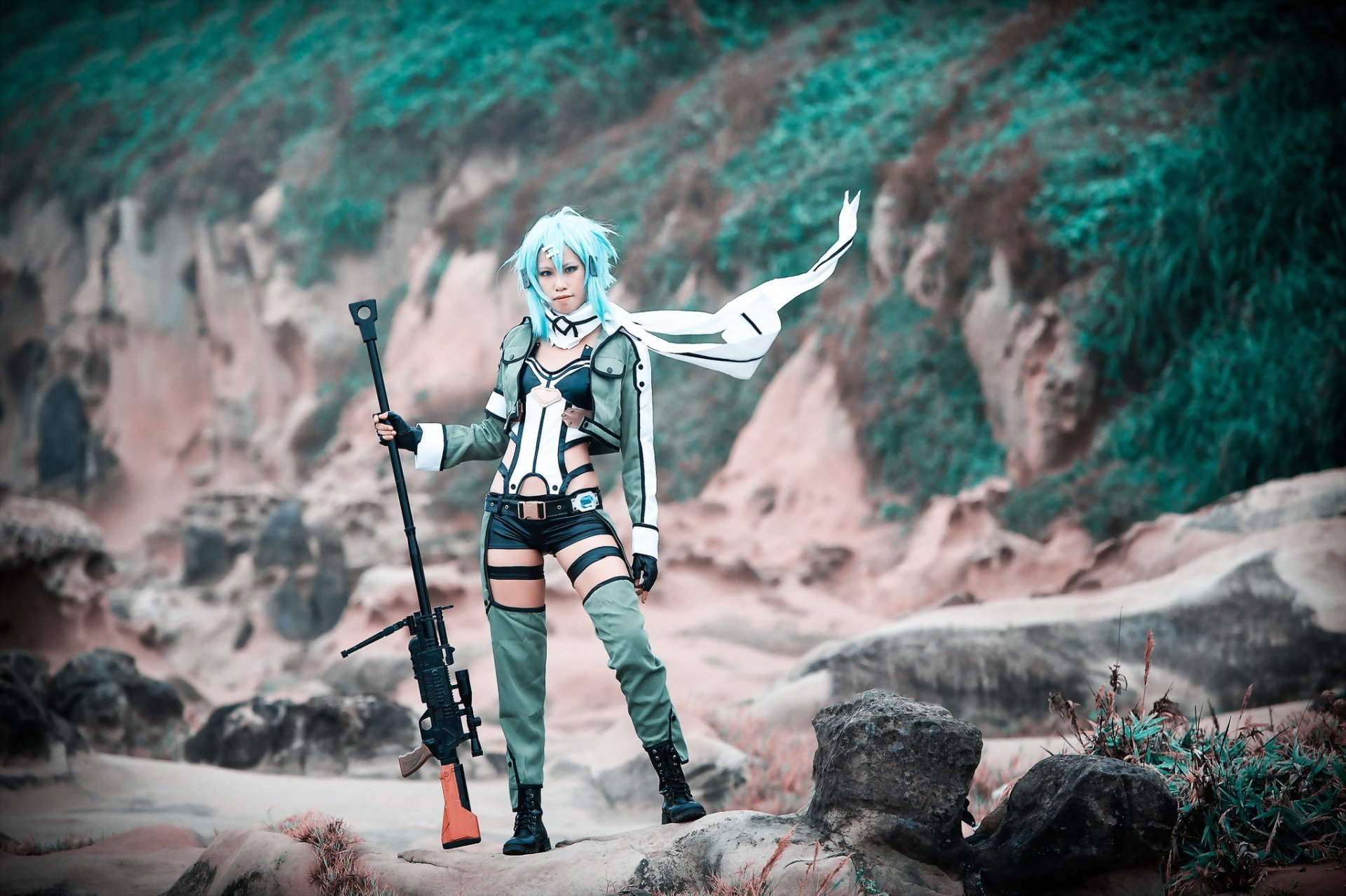 HD desktop wallpaper featuring a woman cosplaying Shino Asada from Sword Art Online, standing confidently in a rocky outdoor setting with a sniper rifle.
