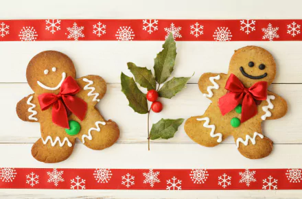 Two smiling gingerbread cookies with red bows flank a sprig of holly on a white wooden surface, bordered by festive red and white snowflake patterns, evoking Christmas holiday cheer.