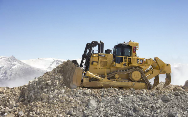 4K Ultra HD image of a yellow Caterpillar Inc. bulldozer moving earth on a rocky terrain with snow-capped mountains under a clear blue sky.