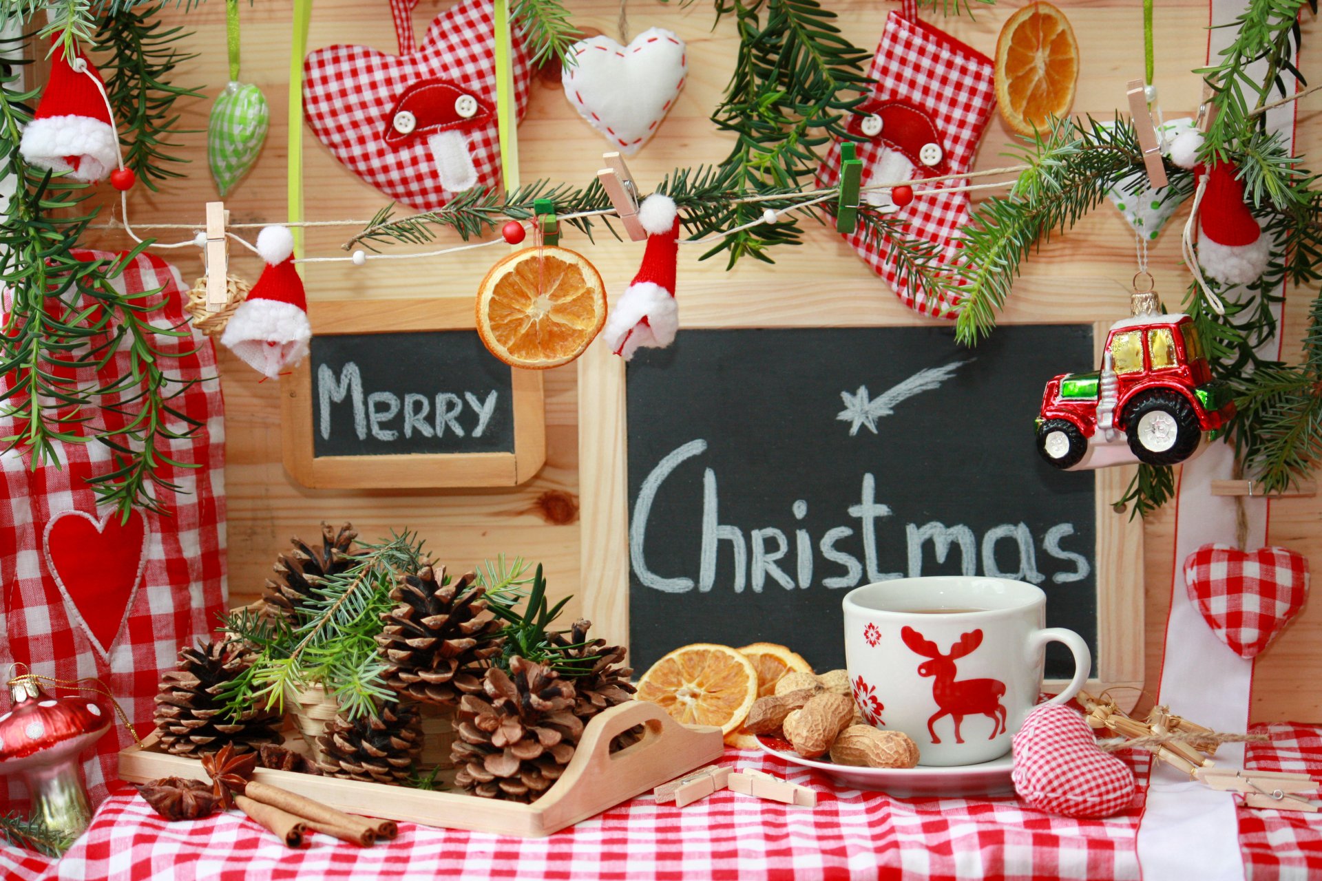Cozy holiday scene with Christmas ornaments, pine cones, a festive mug, and chalkboards reading Merry and Christmas on a red checkered cloth background.