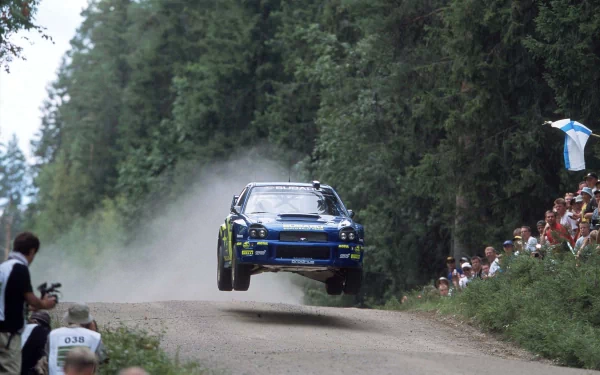HD PC desktop wallpaper of a rallye vehicle airborne on a dirt road, surrounded by spectators and dense forest.