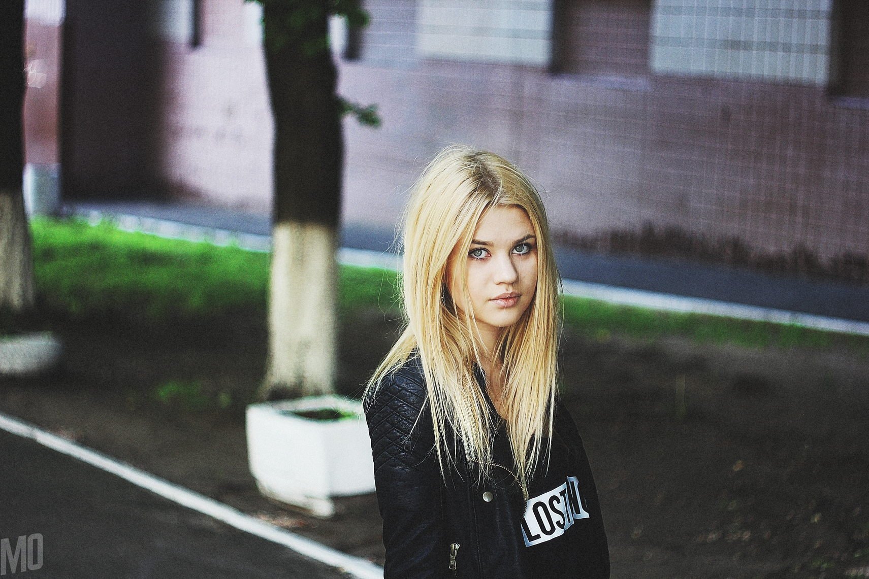 HD desktop wallpaper featuring a blonde female model wearing a black jacket, standing outdoors near trees and a brick building background.